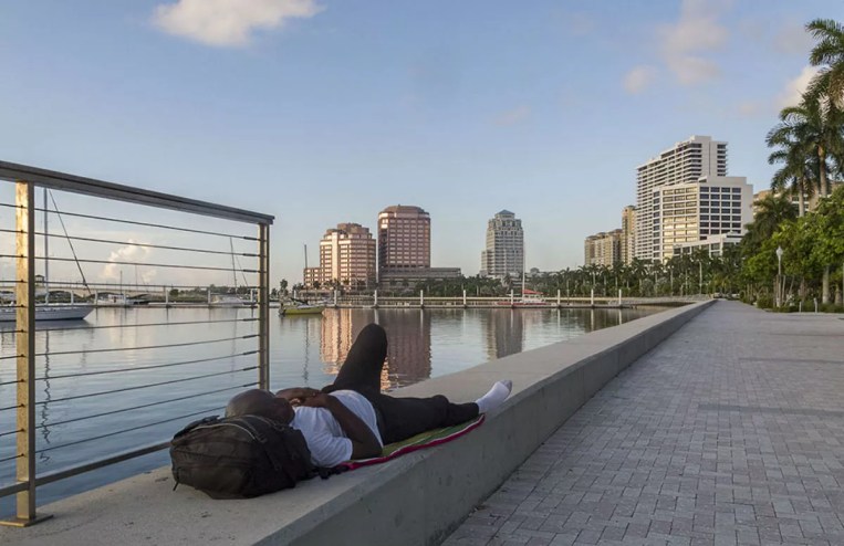 A person reclining on a seawall with their head resting on a backpack.