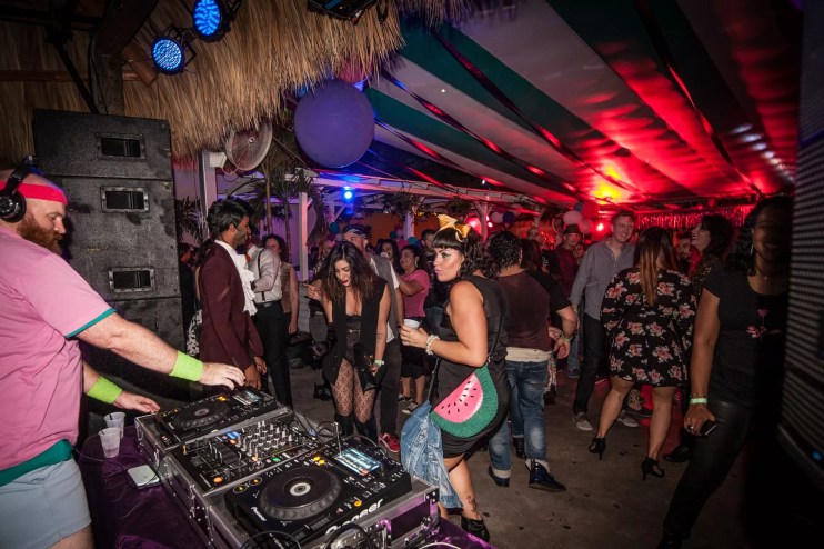 a DJ spins records behind the booth as people dance in from of him