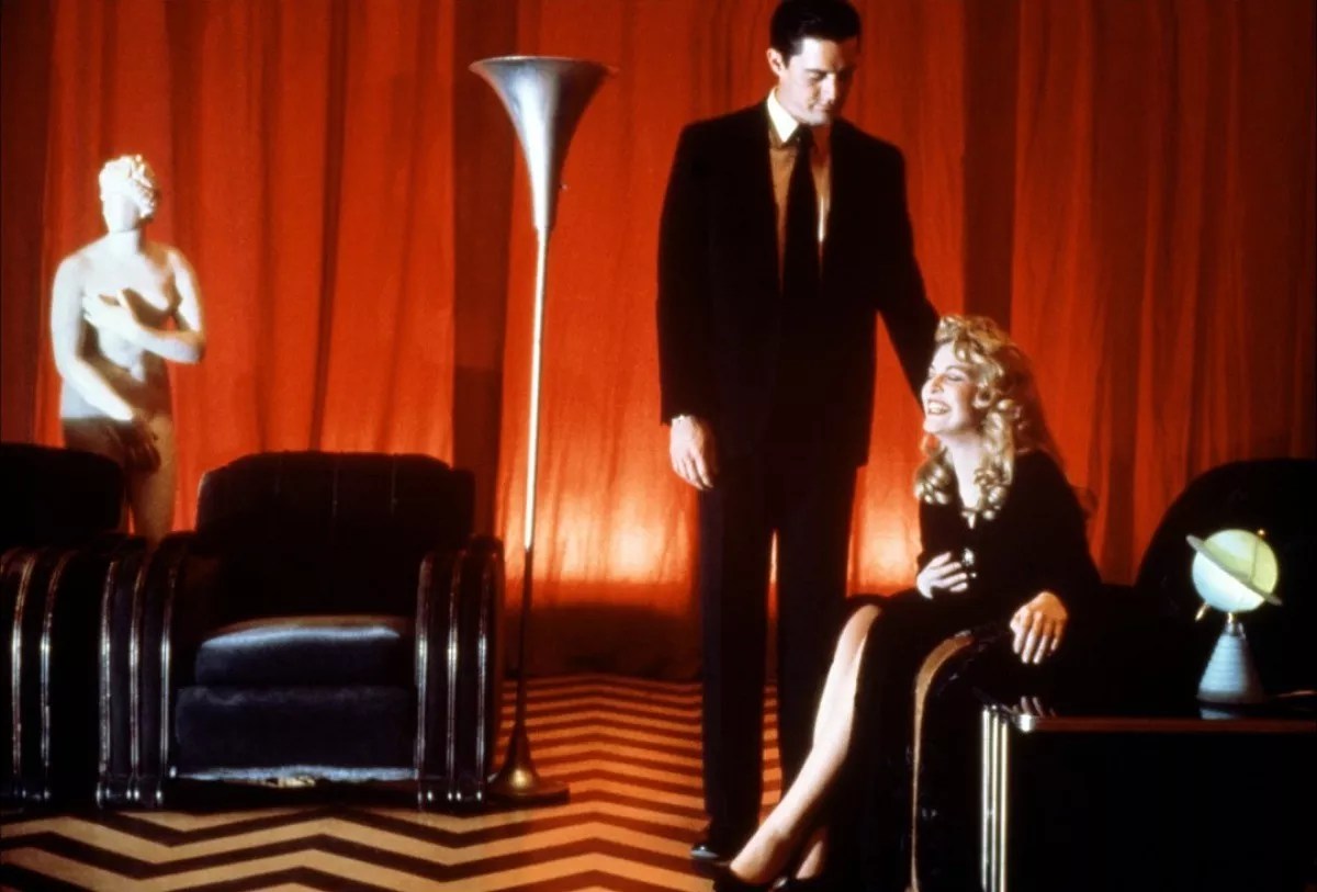 Two characters stand in a room with red lighting and red curtains. The man is standing in a black suit. The woman is sitting in a long black dress. Decor surrounds them, including black leather chairs, a marble statue, and a light-up globe.