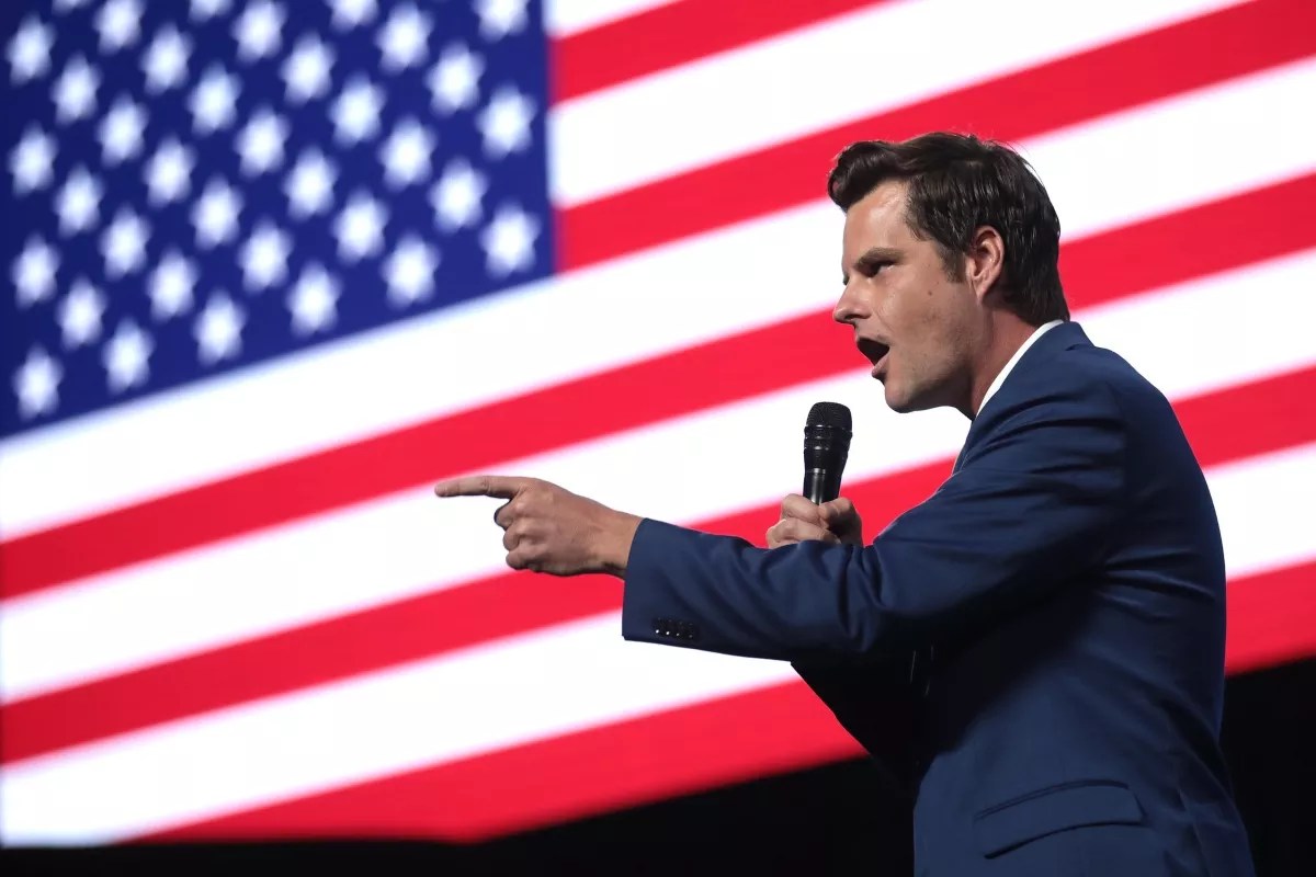 photo of matt gaetz holding a microphone and pointing, presumably to a crowd, against a large american flag backdrop