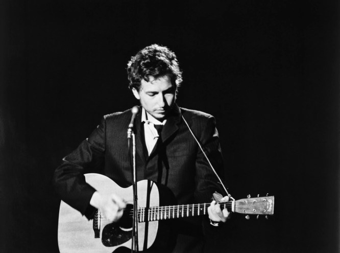 Black and white photo of Bob Dylan on stage in 1969, singing and strumming an acoustic guitar