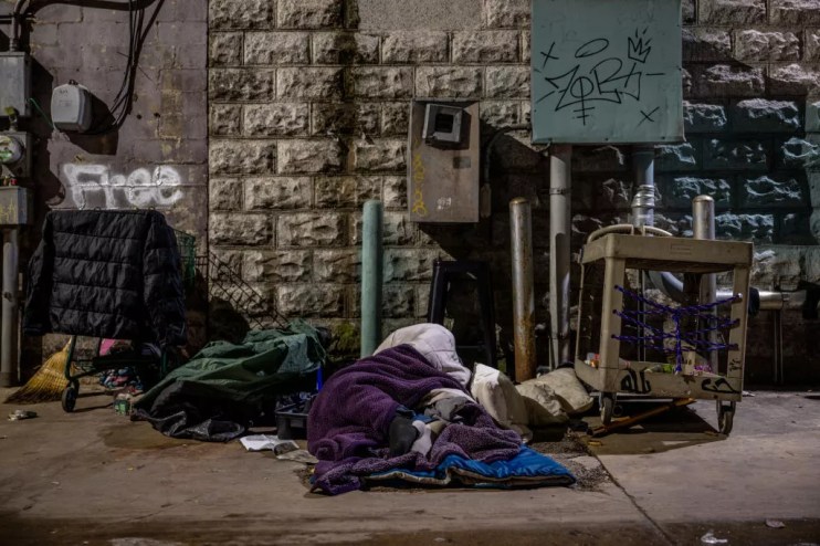 A homeless person lies wrapped in a blanket on the concrete in front of a wall with the word, "Free," painted on it