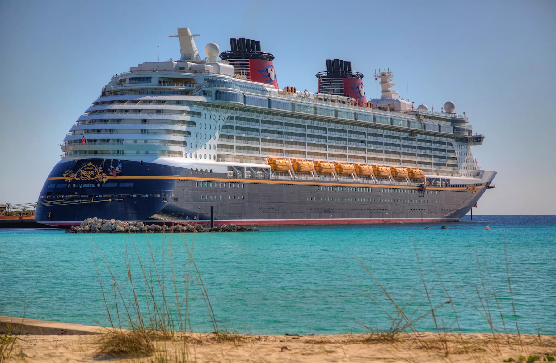 The Disney Dream cruise ship –– a massive vessel colored navy, white, and red –– arrives to shore in turquoise blue waters Castaway Cay, Bahamas.