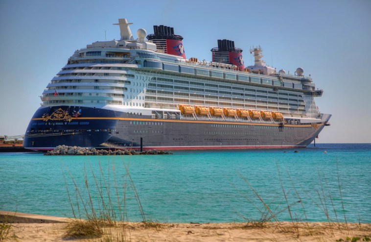 The Disney Dream cruise ship –– a massive vessel colored navy, white, and red –– arrives to shore in turquoise blue waters Castaway Cay, Bahamas.