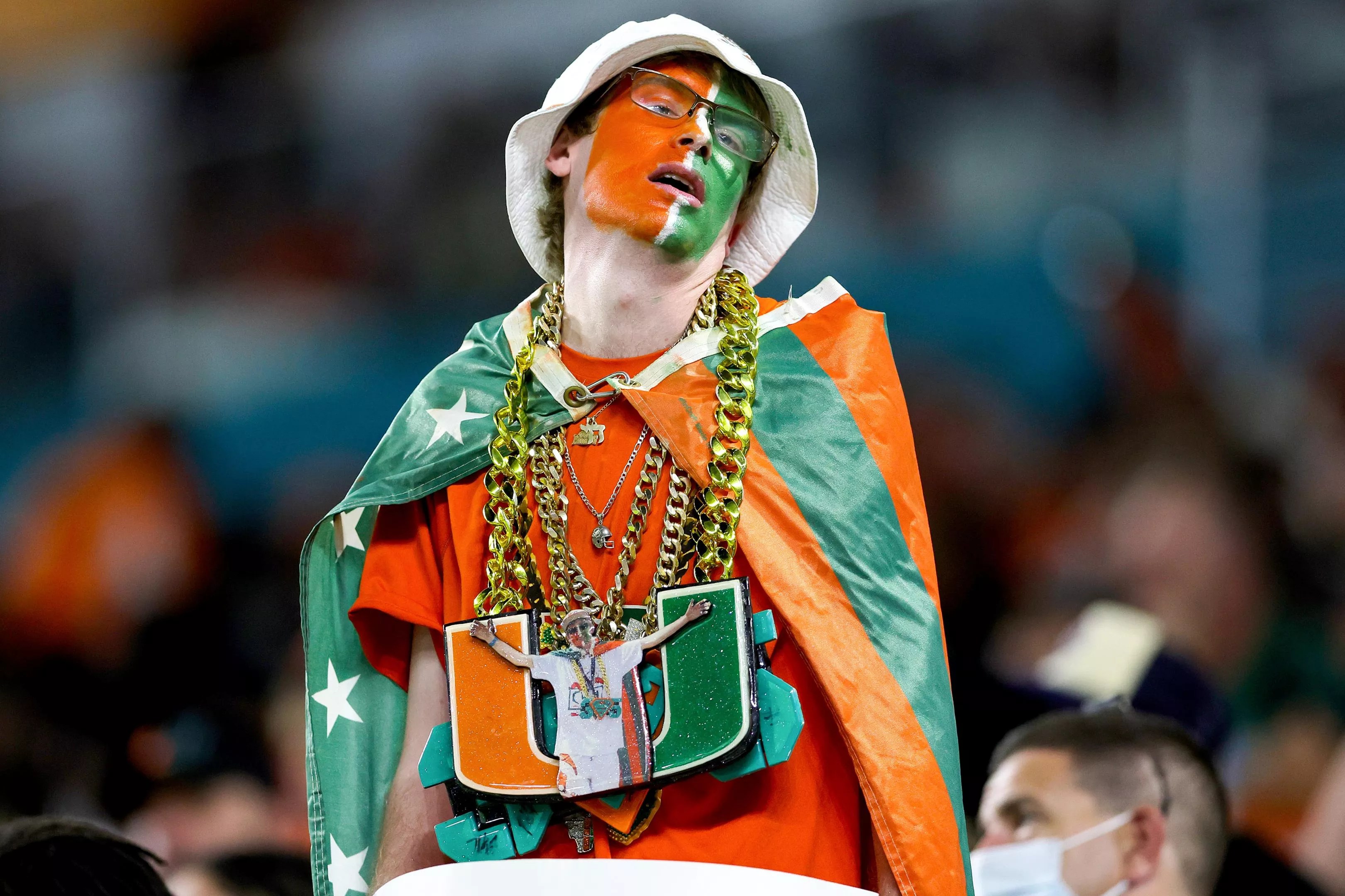 A Miami Hurricanes fanatic with his face painted is wearing gold chains and some kind of cape at a college football game