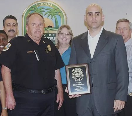 A man with a shaved head poses in plainclothes with fellow police officers, holding an "Officer of the Month" award