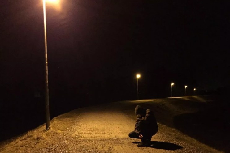 Skee Mask, face obscured, crouching down a road with a street lamp shining overhead