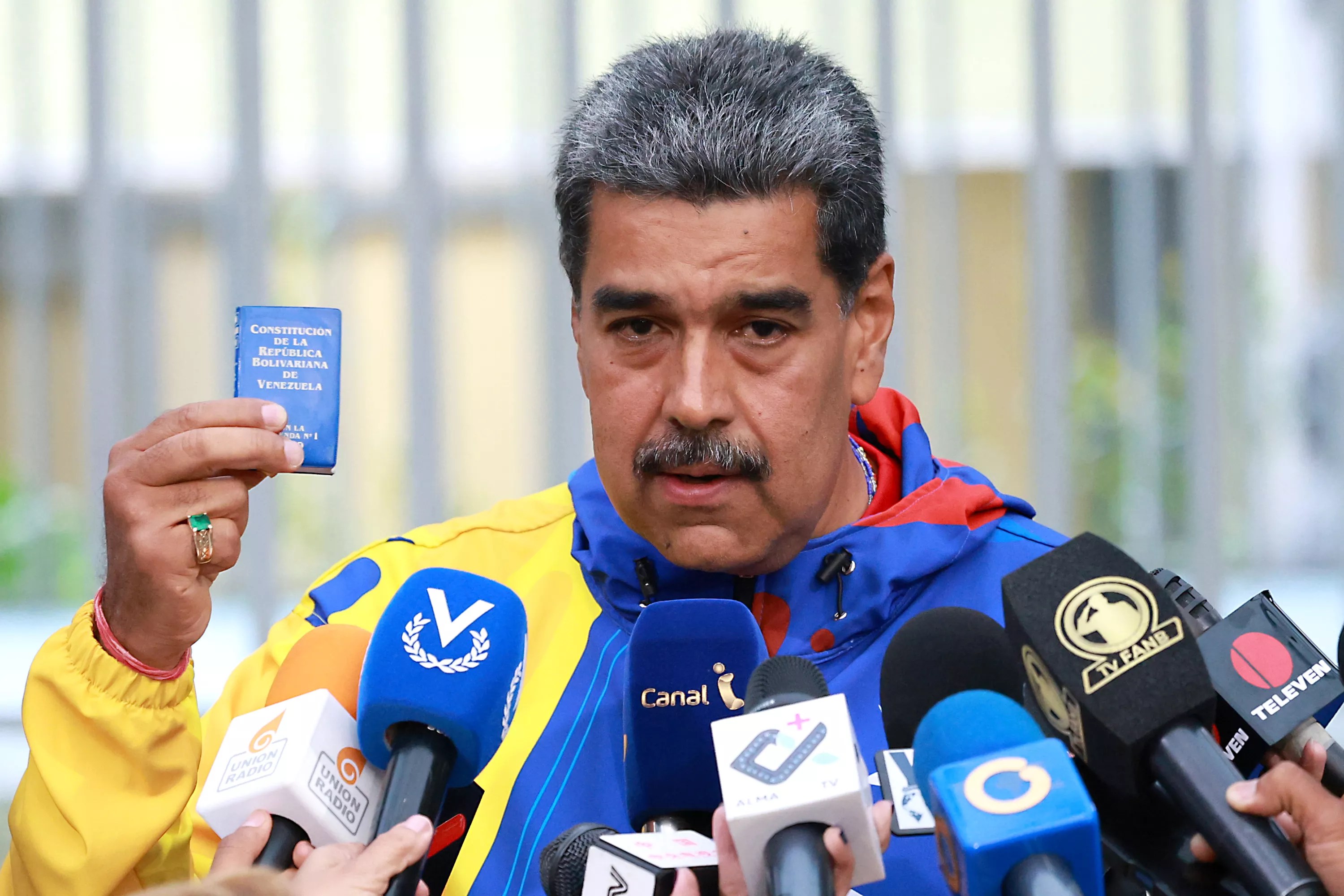 Mustachioed man in front of microphones, holding a teensy copy of the Venezuelan constitution.