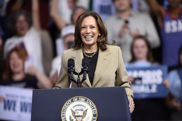 photo of Kamala Harris smiling widely while standing at a lectern bearing the presidential seal, with a crowd of supporters in the background