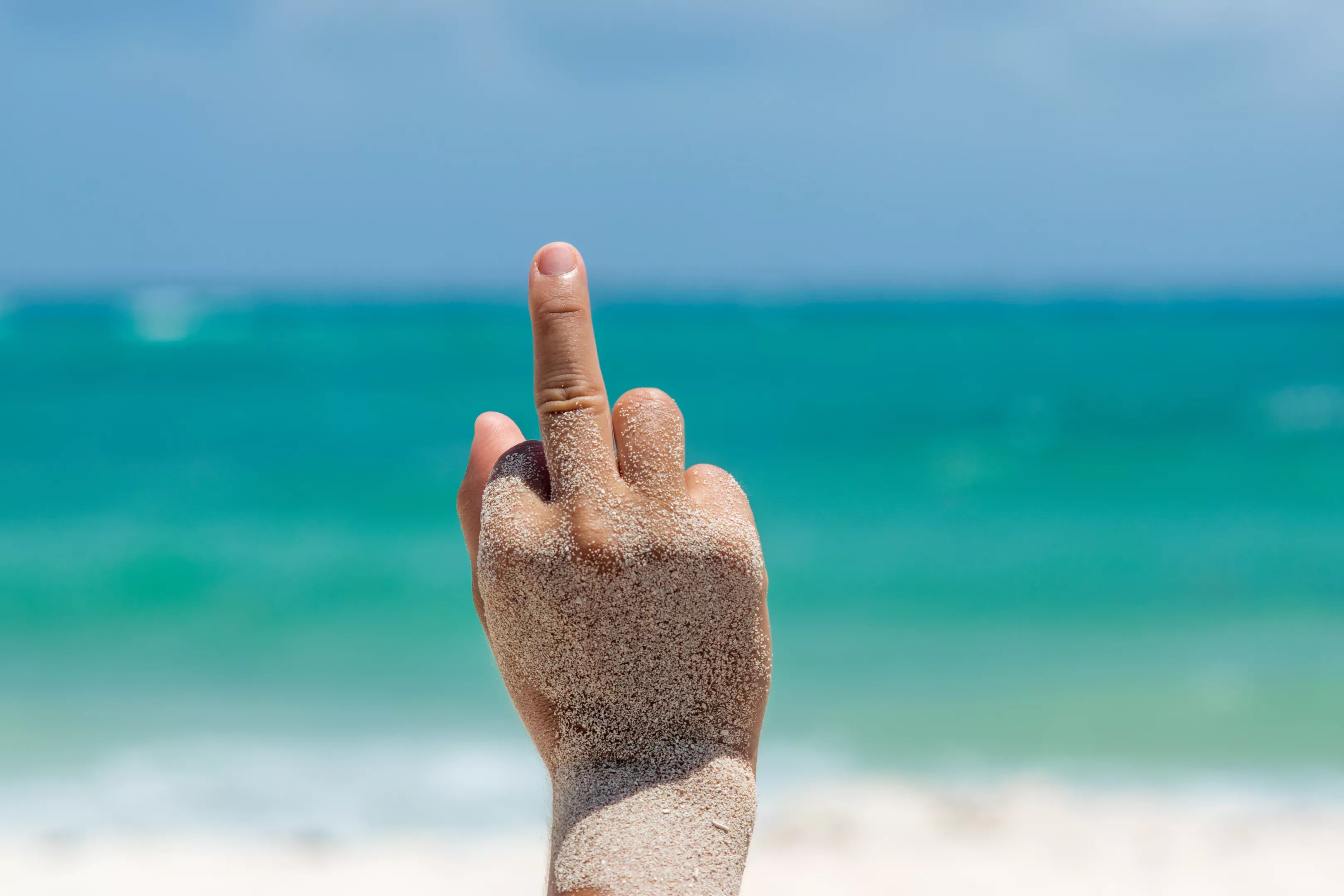 Female hand covered with sand performs a Fuck you Sign with her hand in a tropical beach