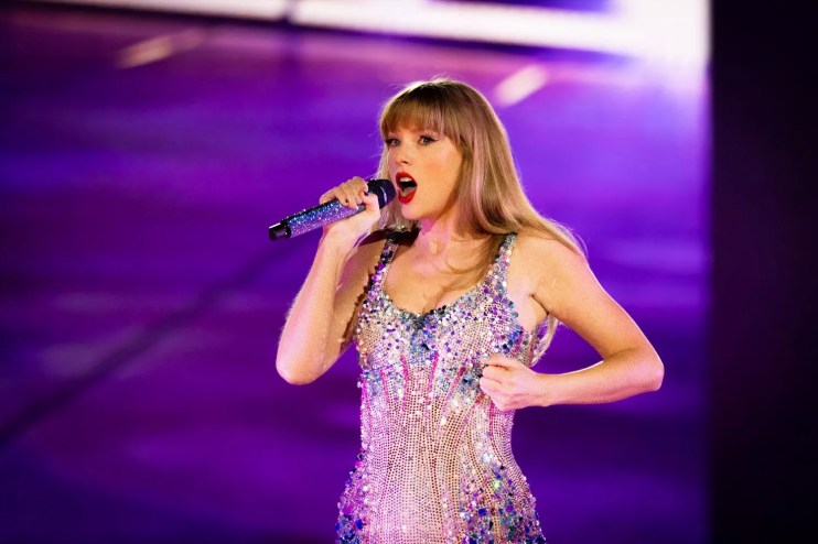 photo of be-sequined taylor swift against a purple backdrop, singing into a bedazzled wireless microphone