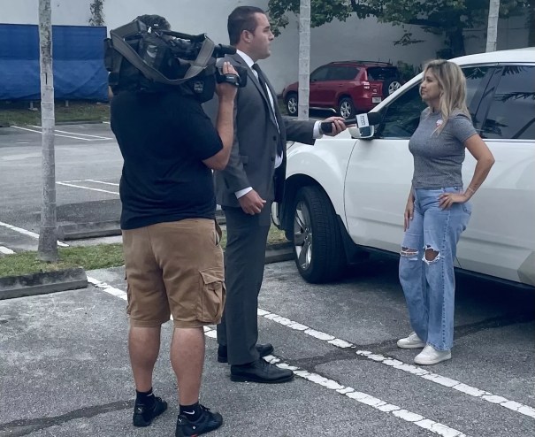 television reporter and cameraman interview a woman standing next to her white car
