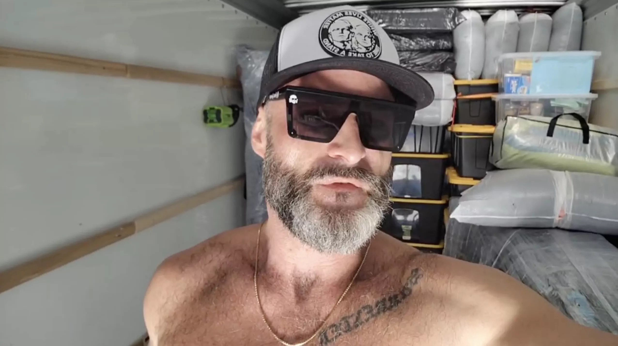 a shirtless Jon Minadeo speaks into the camera inside a loaded U-Haul.