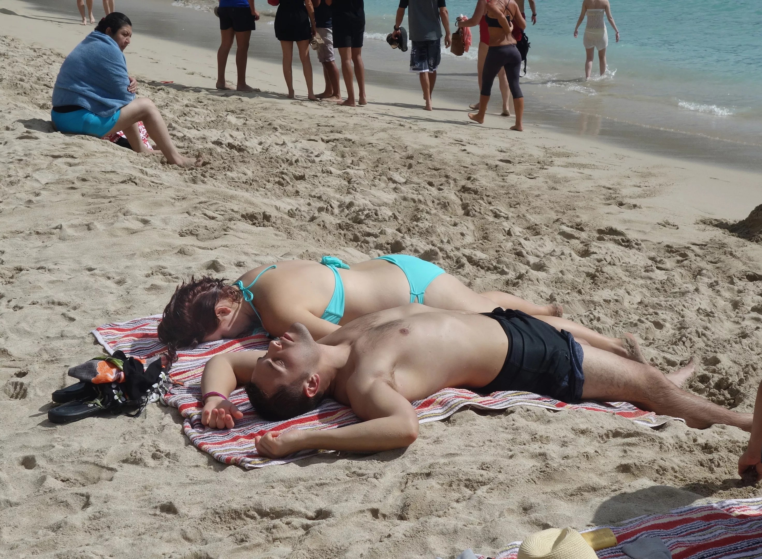 A couple relaxing on sandy shores, lying side by side on a colorful striped towel, soaking up the sun as beachgoers stroll nearby