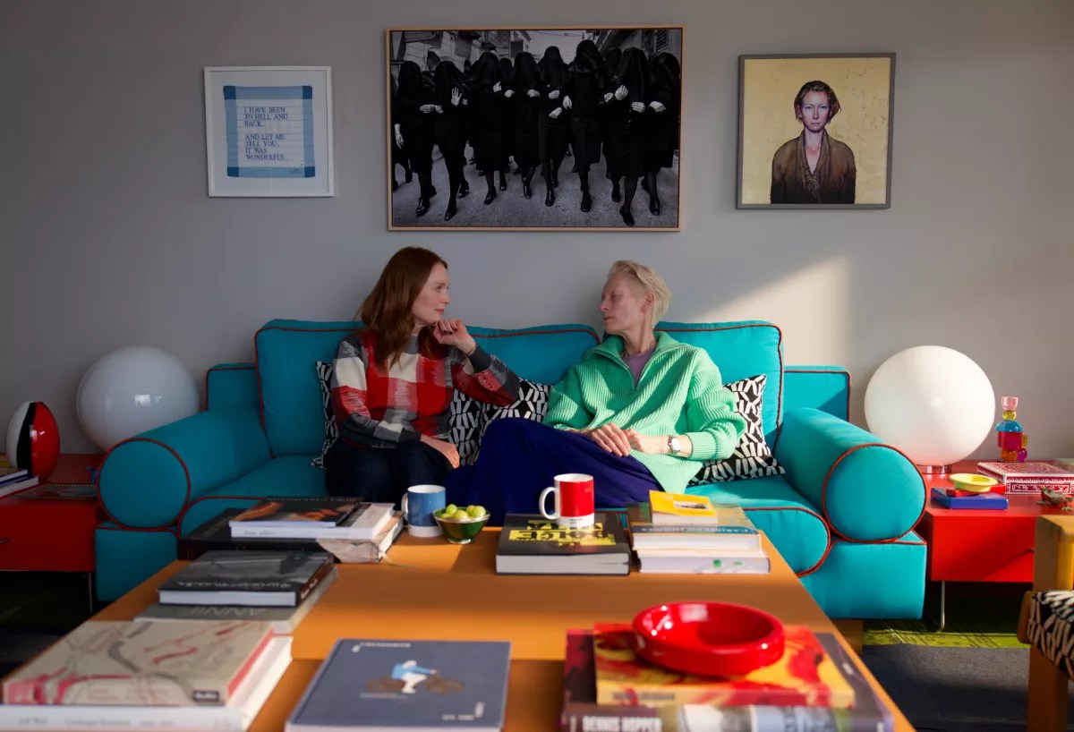 Still of Tilda Swinton and Julianne Moore sitting on a teal couch. Framed photos hang above the couch