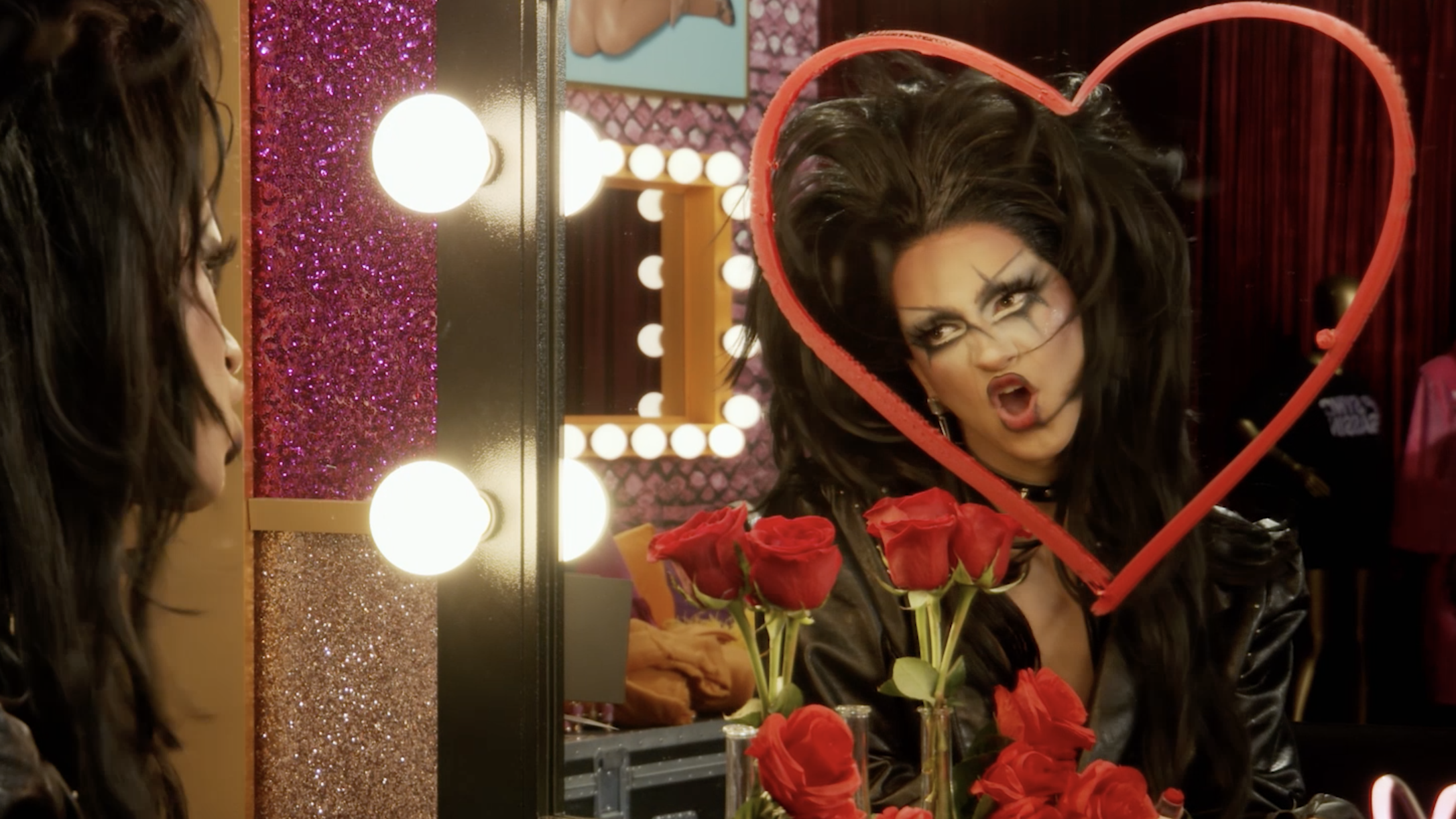 drag race looking at herself in the mirror. a heart is drawn in lipstick on the mirror