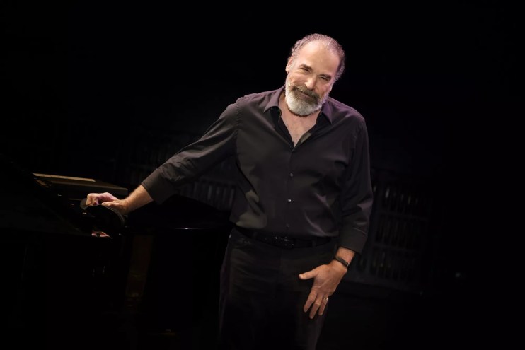 portrait of Mandy Patinkin in a black button-down shirt and pants, leaning on a black piano inside a black box theater