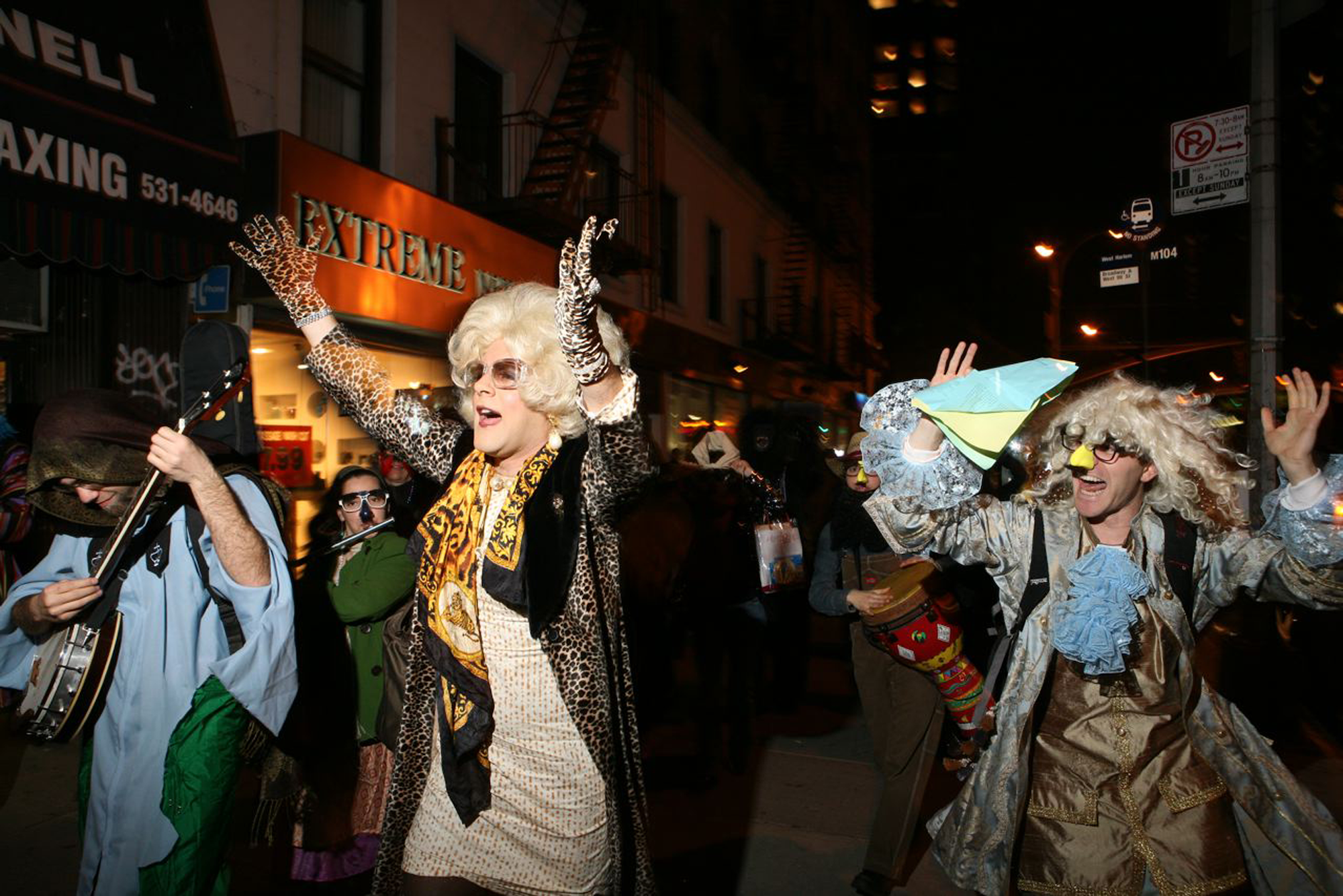 People in costumes dance down the street. A drag queen in a blonde wig and leopard-print gloves and jacket stands in the center.