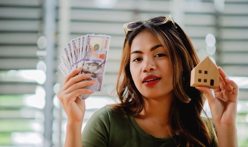 Asian woman coyly posing with $100 bills in one hand and a tiny dollhouse in the other