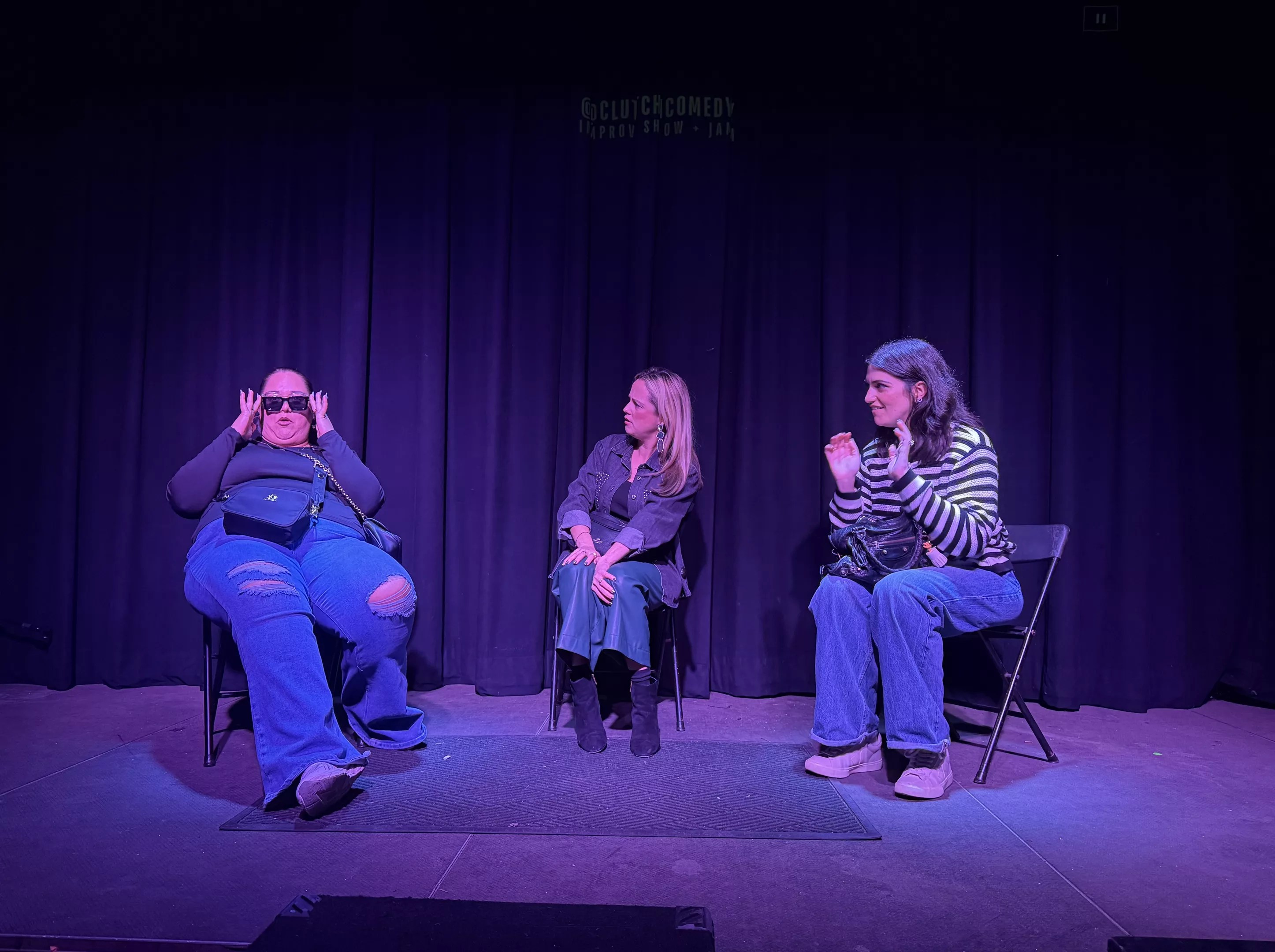 three women sit in chairs in front of a curtain on stage