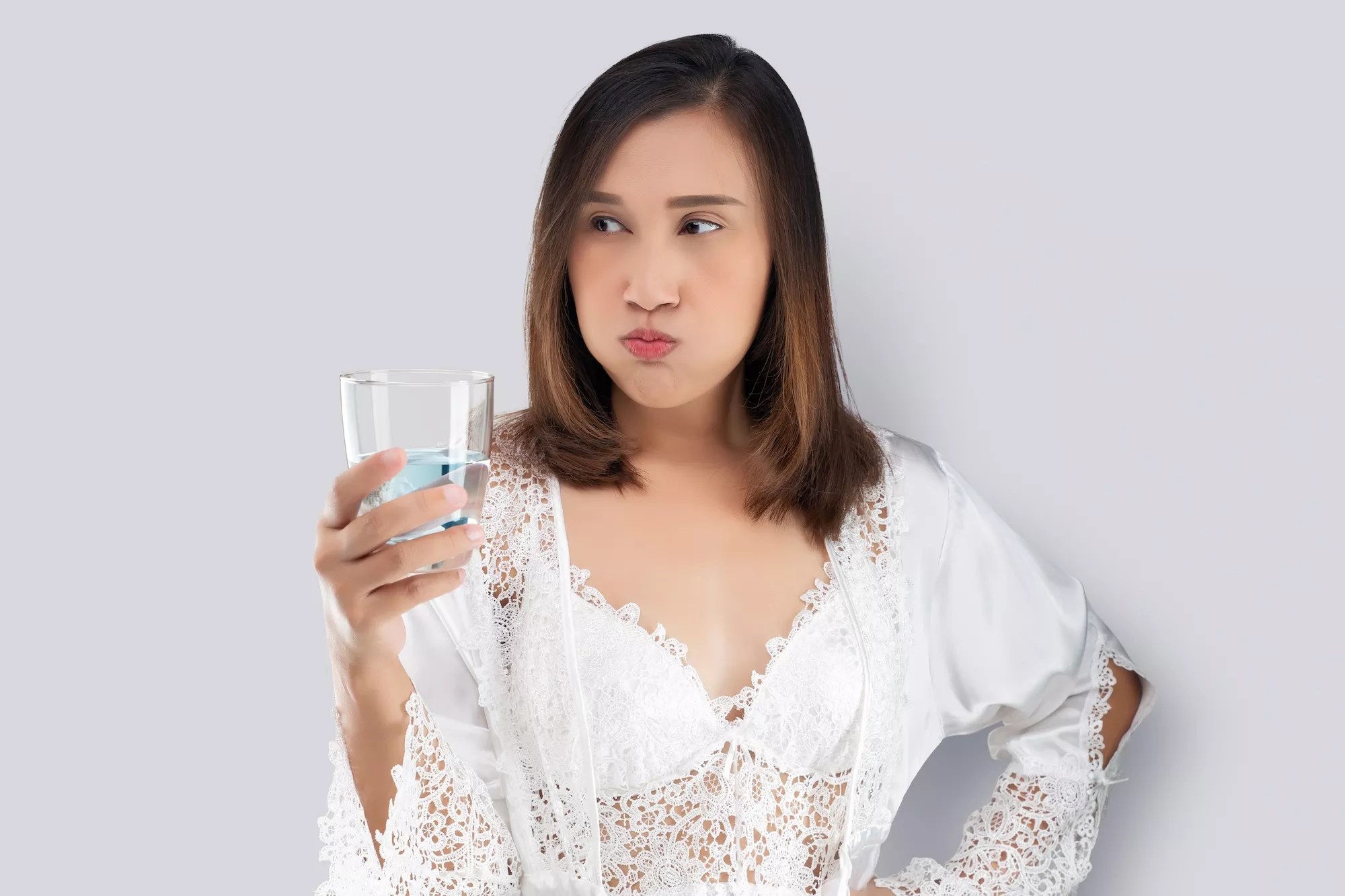 Woman in white silk nightwear and lace robe rinsing with mouthwash against a gray background