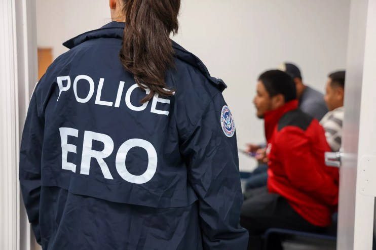 A woman wearing a POLICE "ERO" jacket overlooking three men sitting in chairs blurred in the background.