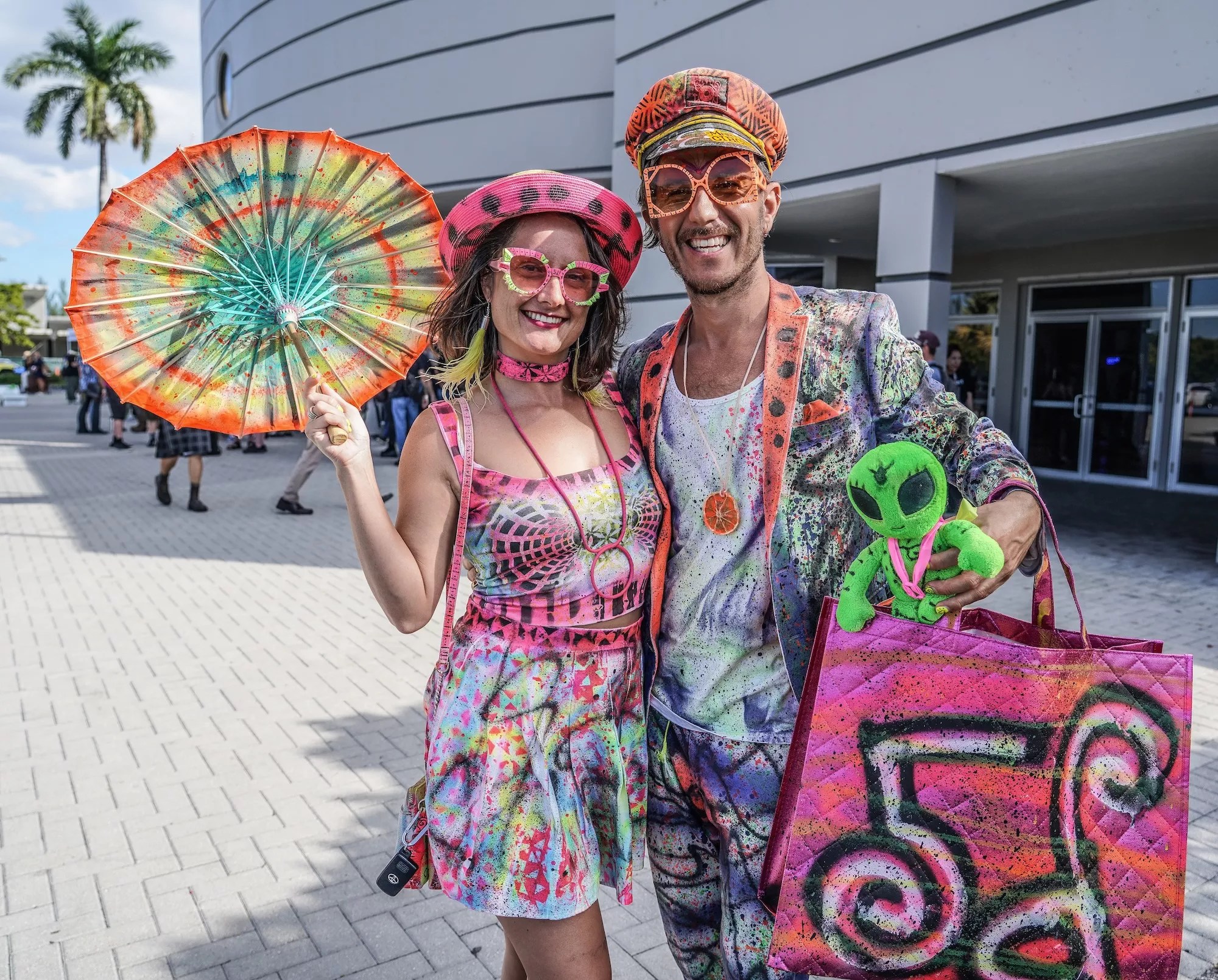 a woman and man in painted colorful clothing poses in front of a convention center. the woman holds a painted parasol