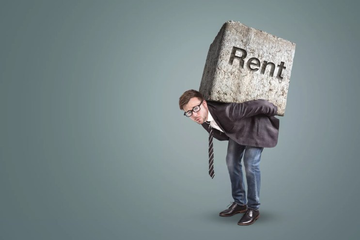 well-dressed hunched man with what looks to be a very heavy concrete block on his back with the word "Rent" etched into it  (Conceptual image of a man burdened by high rent)