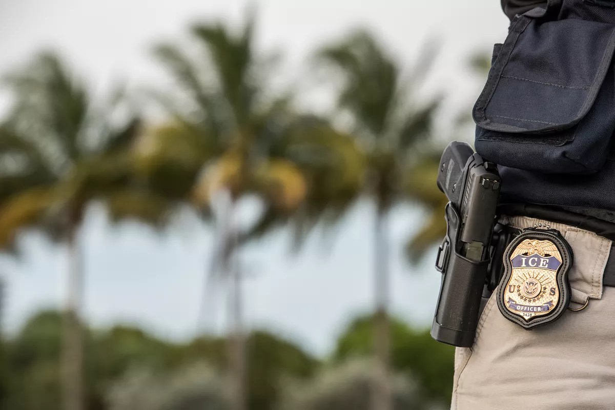 An immigration officer with an ICE badge next to palm trees.