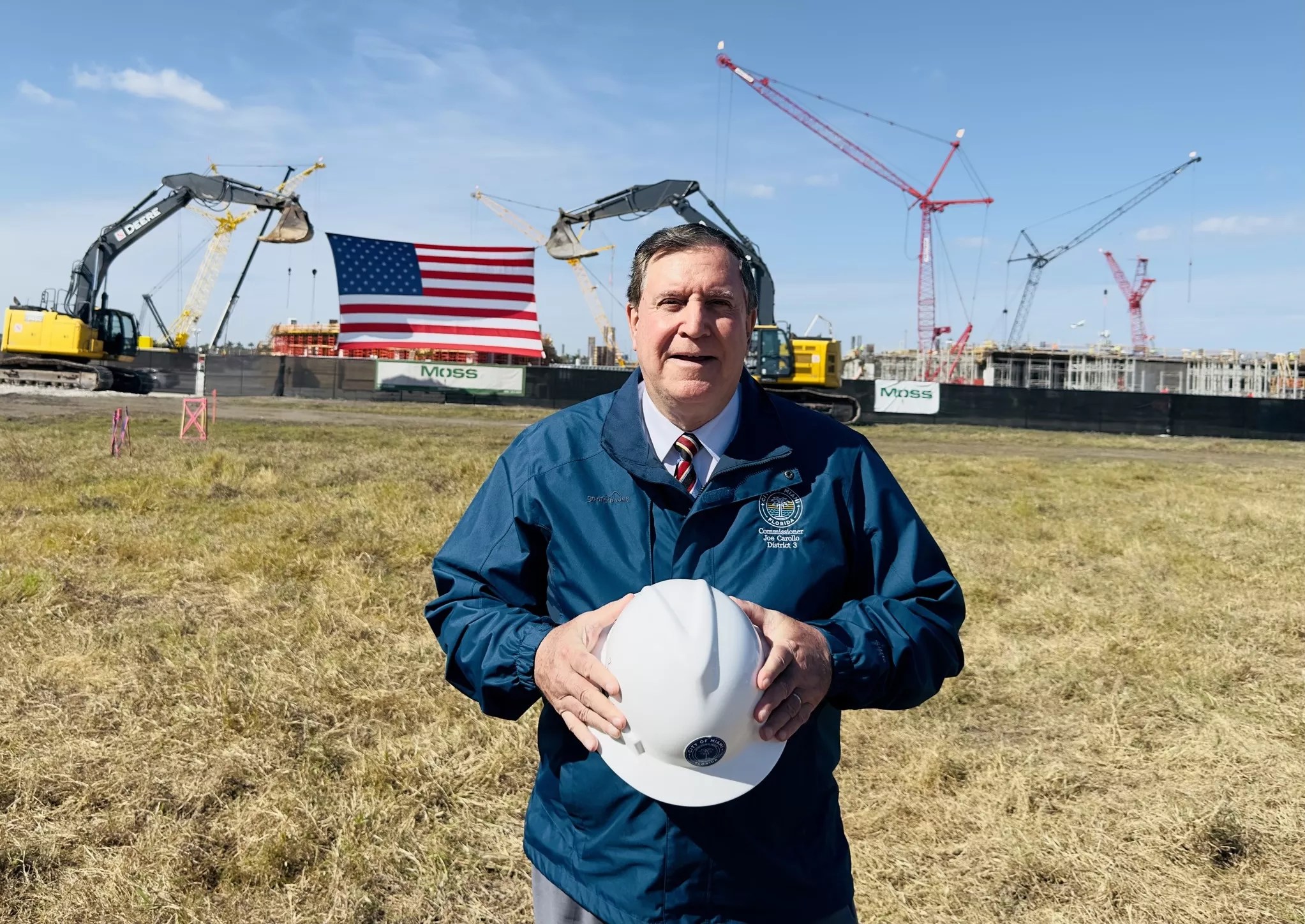 photo of Miami City Commissioner Joe Carollo, wearing a jacket and holding a hardhat in a vacant field with cranes and earth moving machinery in the background