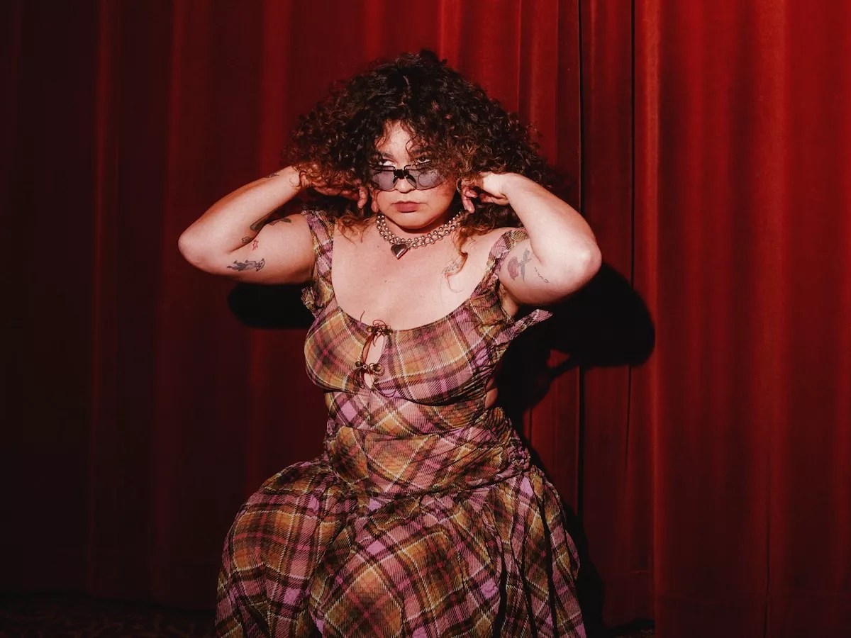 color photo of singer songwriter Remi Wolf, bespectacled, clad in a low-cut plaid dress, and showily primping her hair