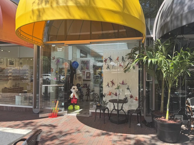 Miami Art Gallery Gift Shop glass storefront, which has a Mickey Mouse sculpture at its entrance, along with a yellow awning.
