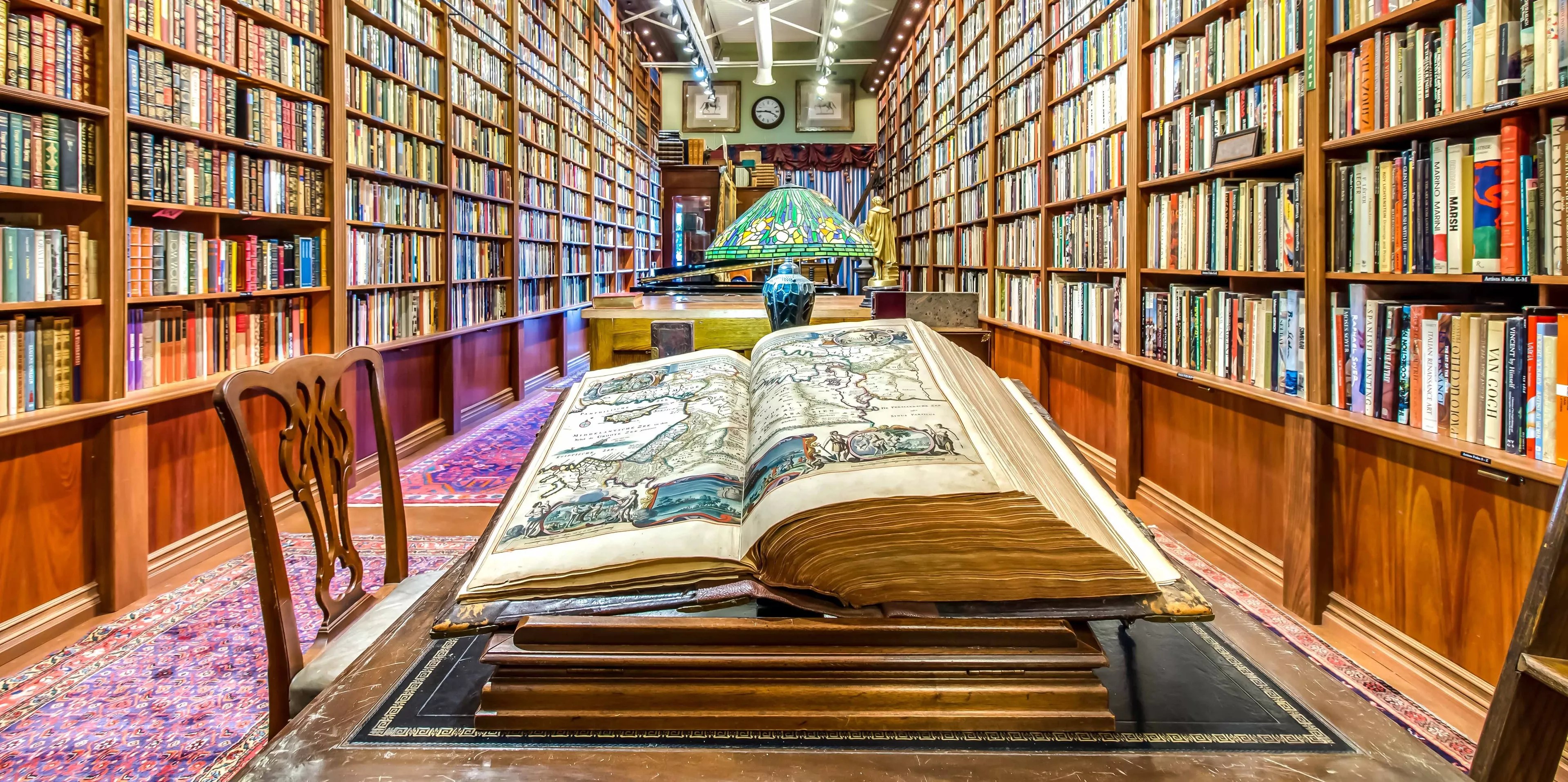 An open book on a table shows a map. Behind it are an antique lamp and bookshelves with hundreds of books