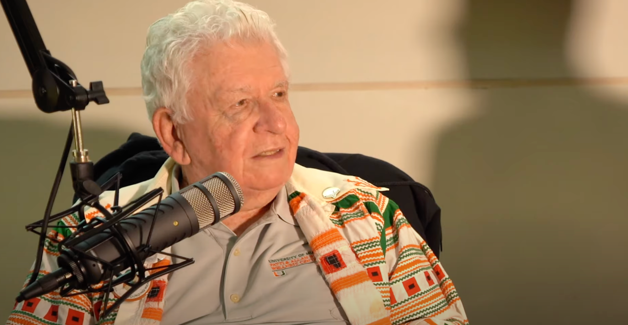 An elderly man with white hair looks to the side in front of a podcast microphone