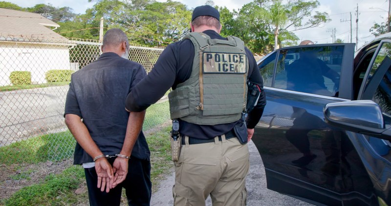 An ICE agent holds a man in handcuffs