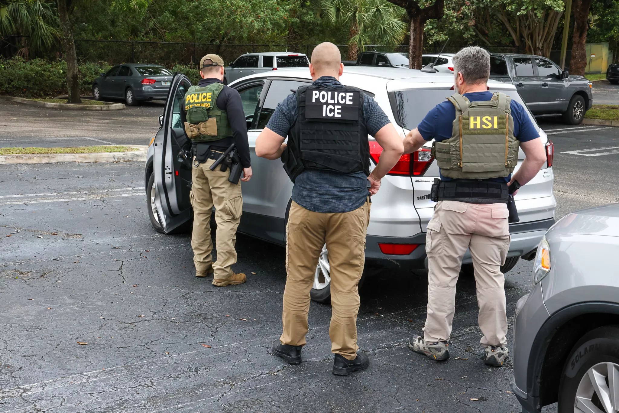 Immigration enforcement officers stand outside a parked vehicle,