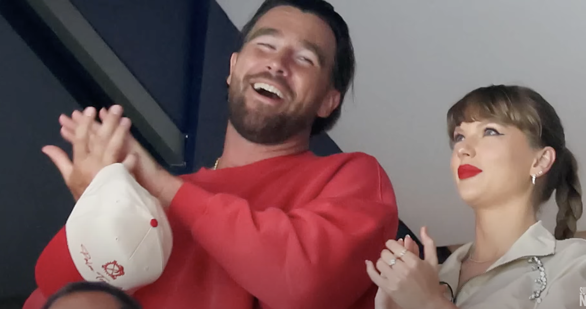 A man in a red sweater and a woman in a khaki top and red lipstick clap and smile