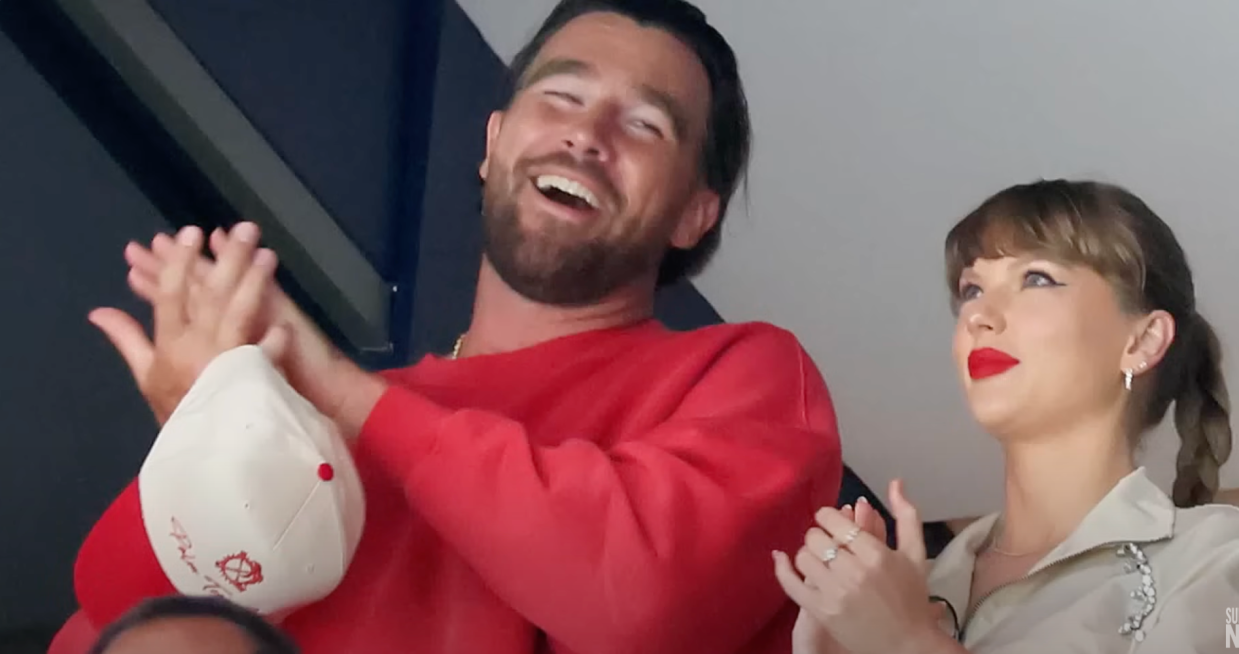 A man in a red sweater and a woman in a khaki top and red lipstick clap and smile