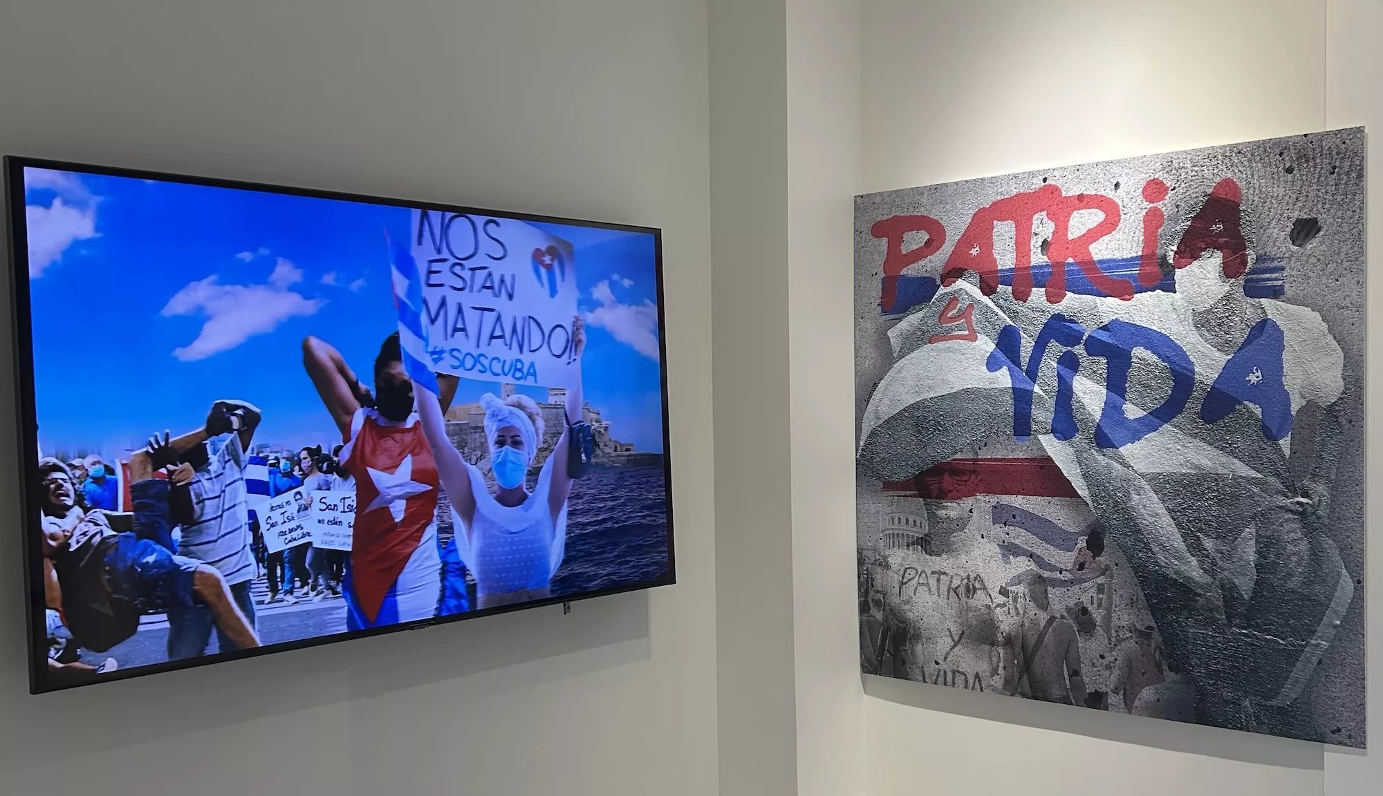 A TV in a museum displays images of Cuban protesters. One holds up a sign reading, "<i>Nos Estan Matando</i> (They're killing us) SOS Cuba." On the right of the TV, a painting displays the slogan, "Patria y Vida (Homeland and Life)"