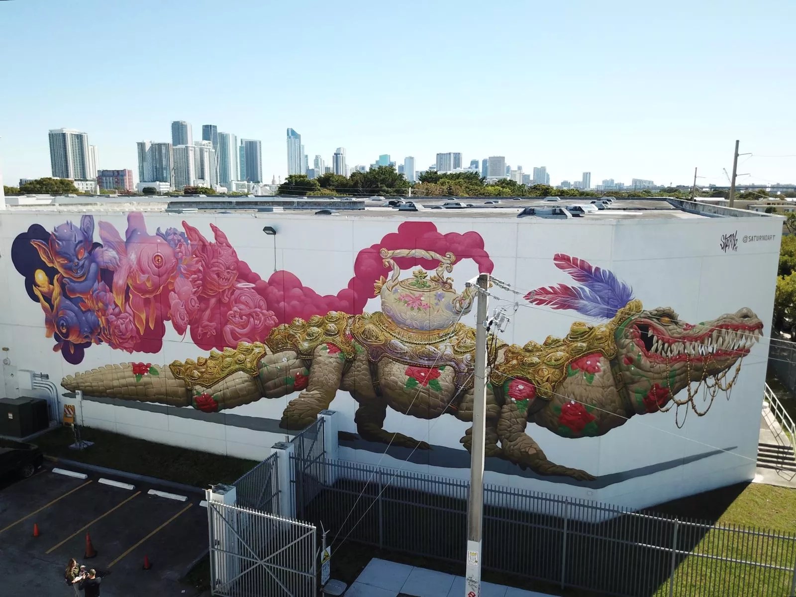 a white building displays a mural with an alligator wearing regal decor, a tea kettle, and feathers