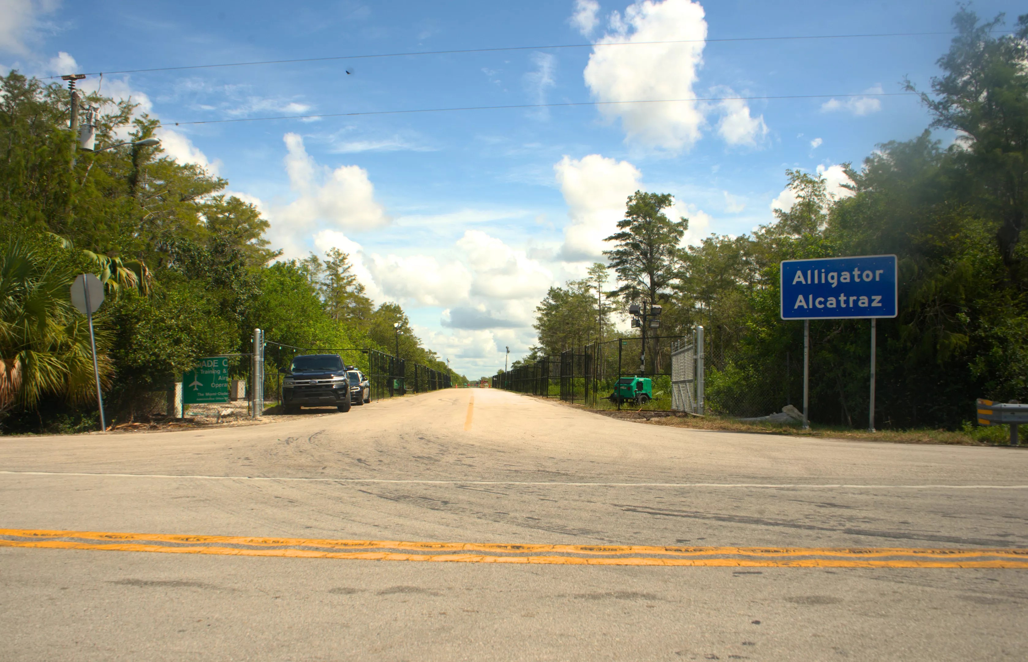 A wide shot of a road with trees on either side, with a blue sign that reads, "Alligator Alcatraz."