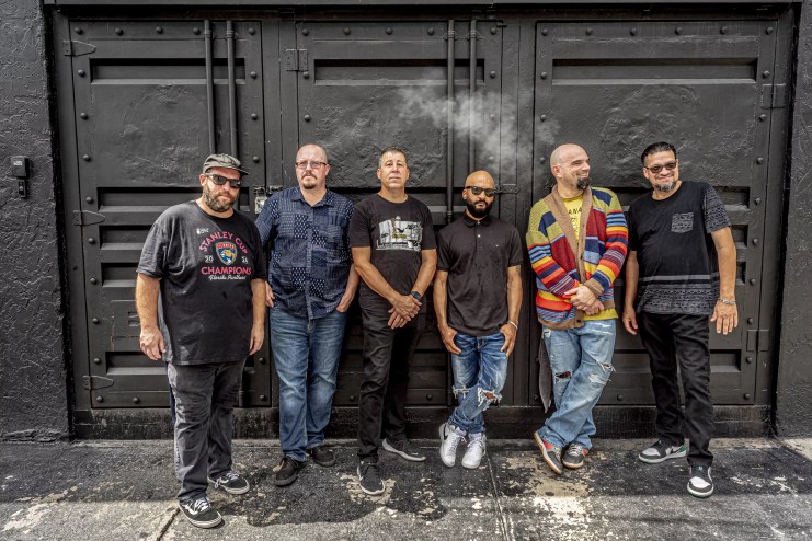 Portrait of a six-piece band leaning on a wall.