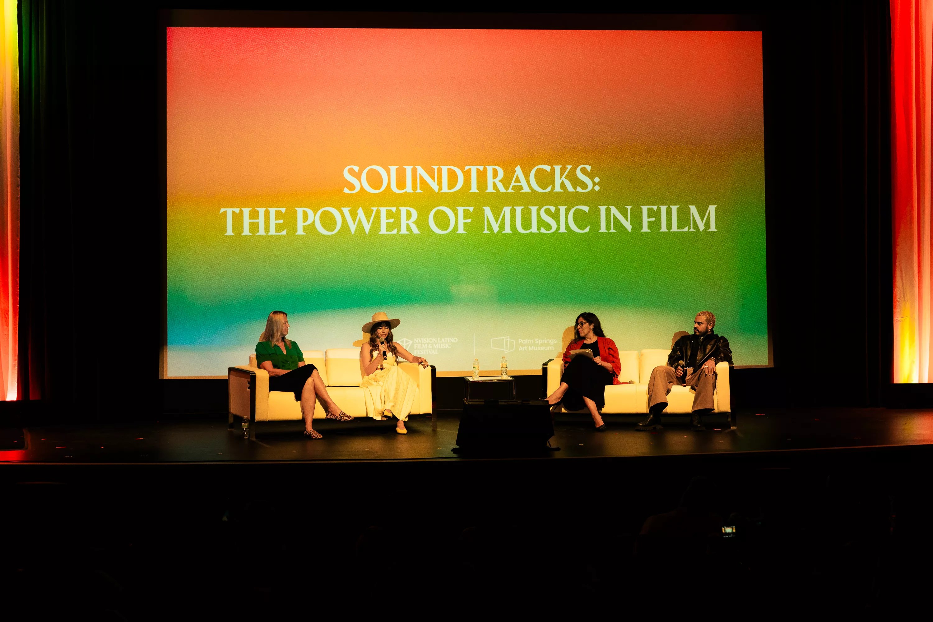 a panel of four people sit on chairs on a stage in front of a projection with the words, "soundtracks: the power of music in film"