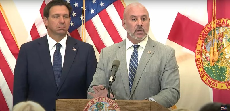 Blaise Ingoglia speaking at a press conference and Ron DeSantis stands to his left with a frown on his face.